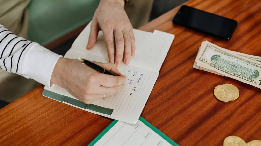 A person writing a budget in a notebook beside dollar bills and a phone on a wooden table