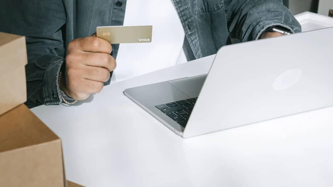 A business owner holds a card beside a laptop and a stack of shipping boxes