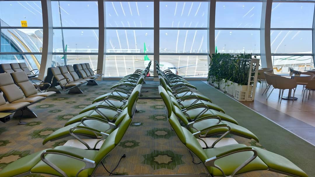 A modern airport seating area with green chairs and large windows looking onto the runway