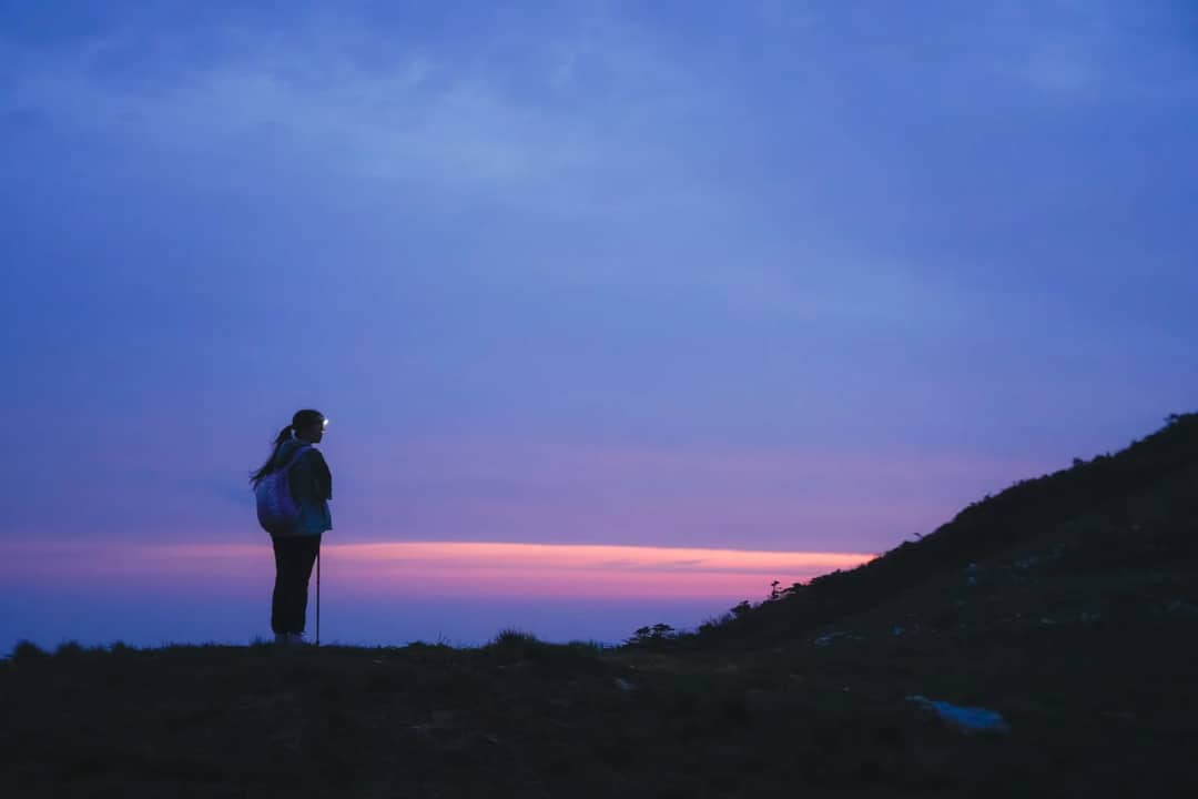 A lone hiker standing at twilight watching the last light fade on the horizon