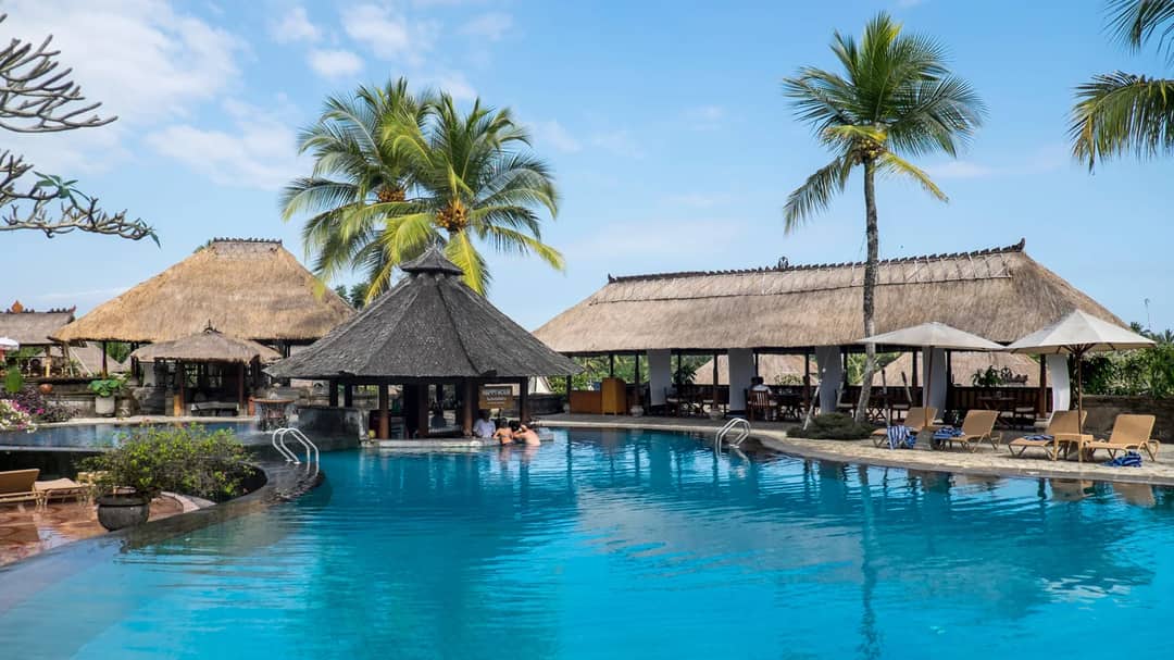 A resort swimming pool with palm trees and thatched pavilions