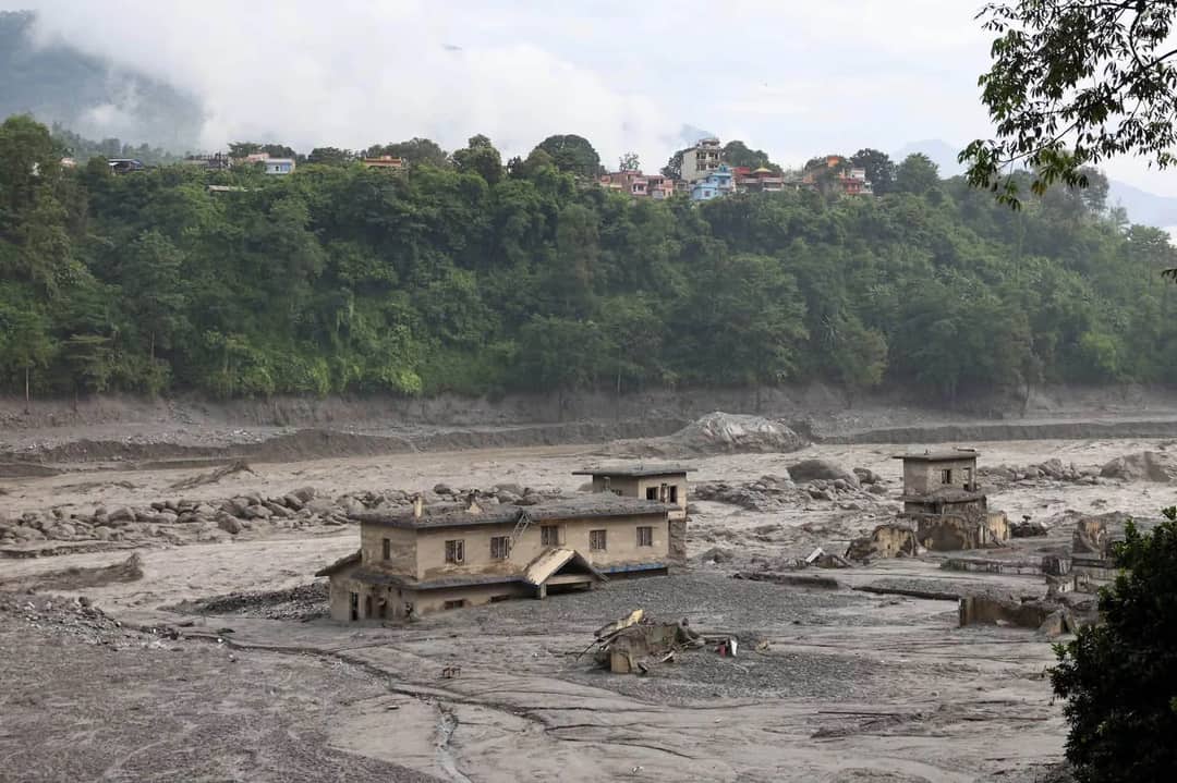 Buildings gutted and half-buried in grey silt on the Trishuli riverbank in Nuwakot after the 2026 flood, with an intact hillside settlement above