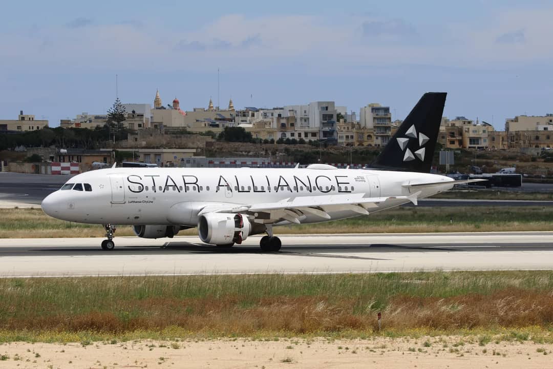 Star Alliance airplane taxiing at Malta's airport, surrounded by cityscape.