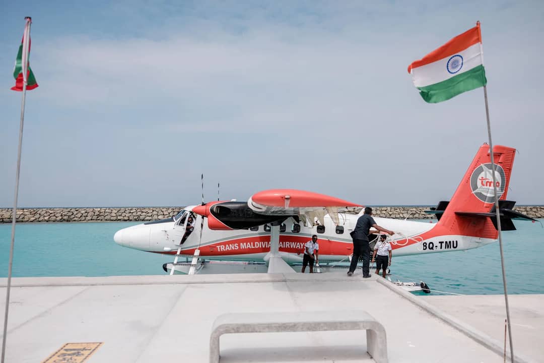 Trans Maldivian Airways seaplane docked at the pier, flanked by national flags under a clear sky.