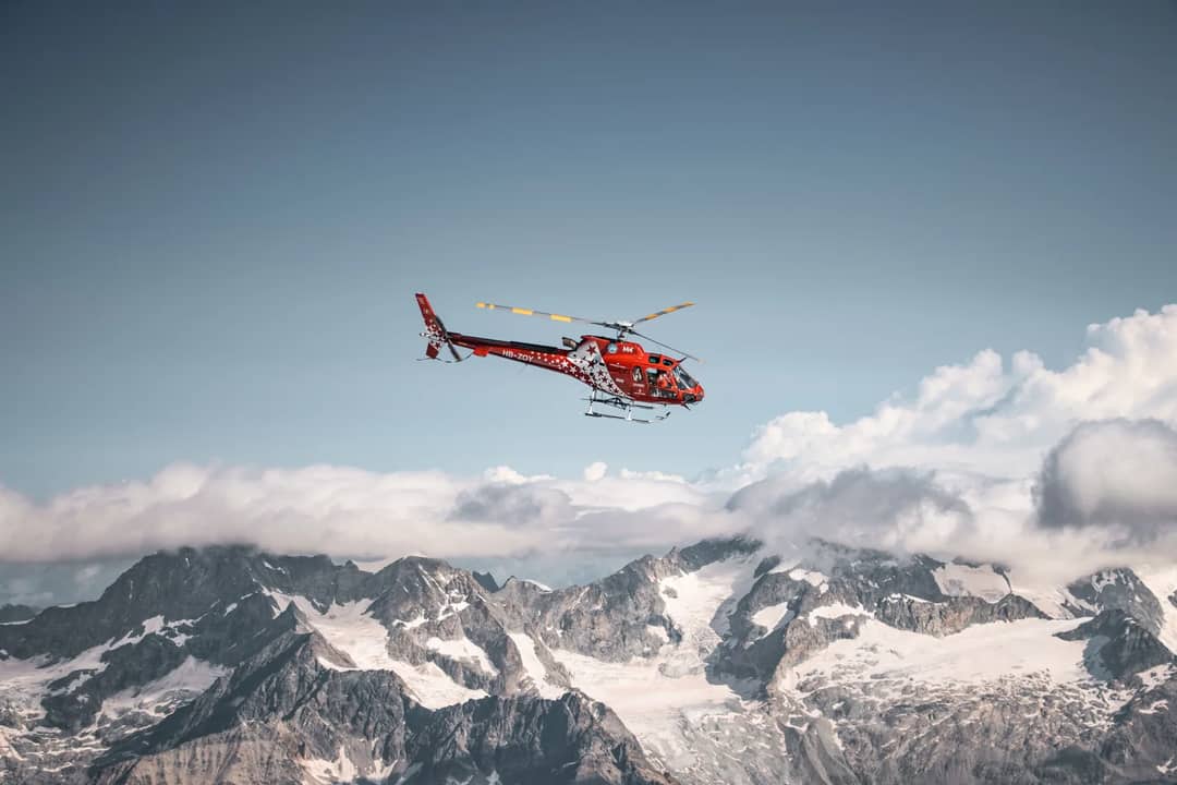 A vibrant red helicopter flies over the snowy peaks of the Swiss Alps, capturing a winter adventure.
