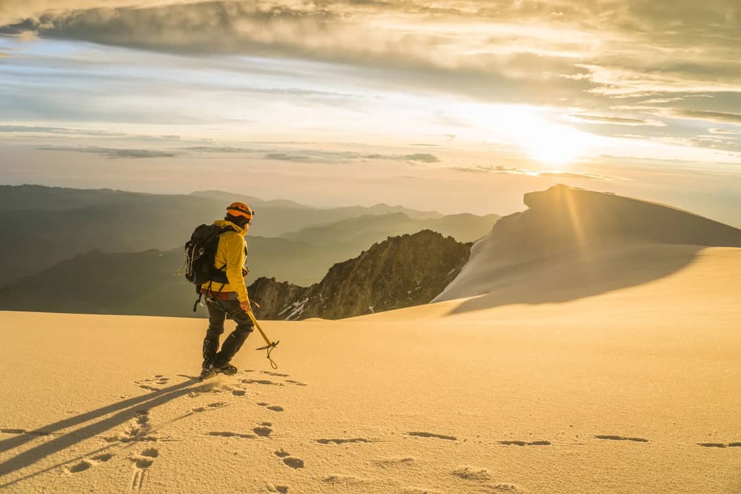 A lone mountaineer walks through a snow-covered mountain range during a golden sunrise.