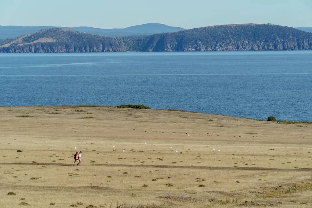 Two hikers traverse the vast landscape of Maria Island, Tasmania, with a serene ocean backdrop.