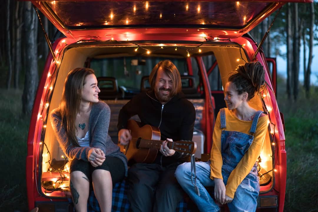 A group of friends in the back of a van, playing guitar and enjoying twilight outdoors.
