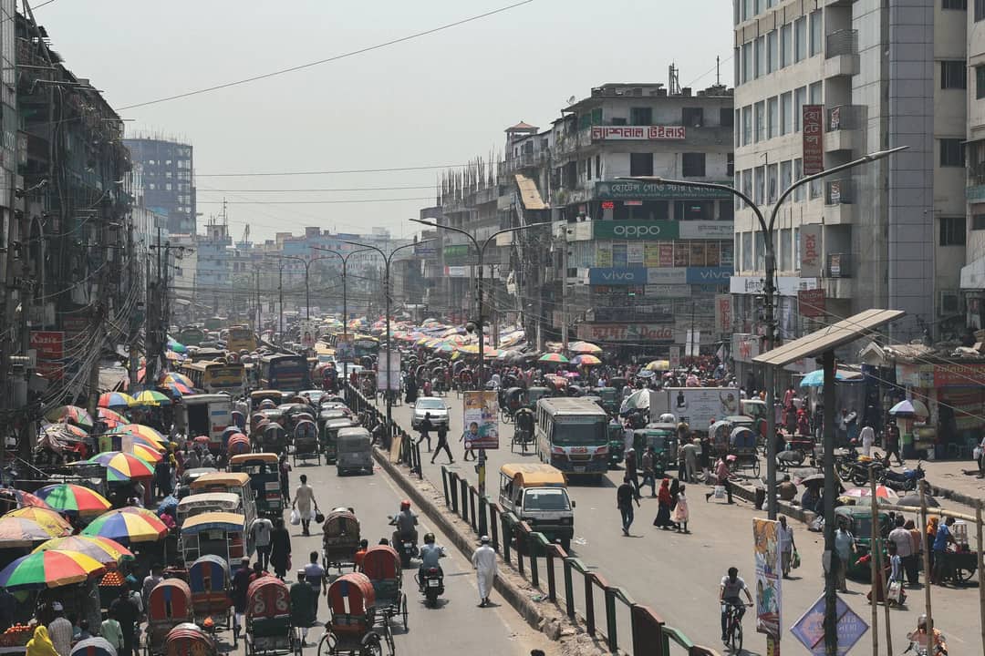 A bustling street scene in Dhaka, Bangladesh with vibrant crowds, vehicles, and colorful umbrellas, viewed from above.