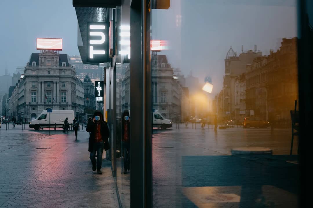 Anonymous people in medical masks walking on Place de Brouckere square near modern building with glass walls in rainy evening during coronavirus in Brussels