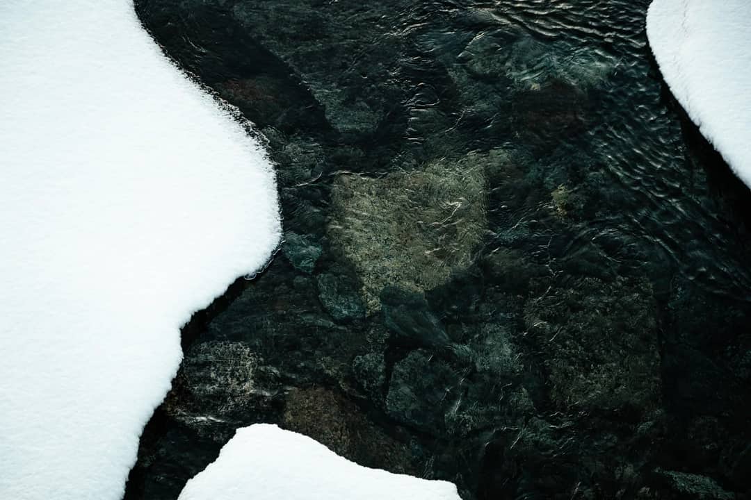 A serene winter stream flows over rocks, framed by untouched snow, capturing nature's tranquility.