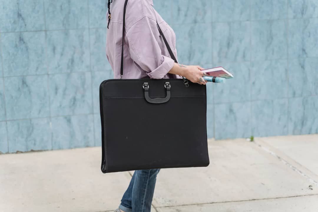 Side view of crop anonymous woman in trendy outfit standing near blue wall in street and holding big black art bag and notebook