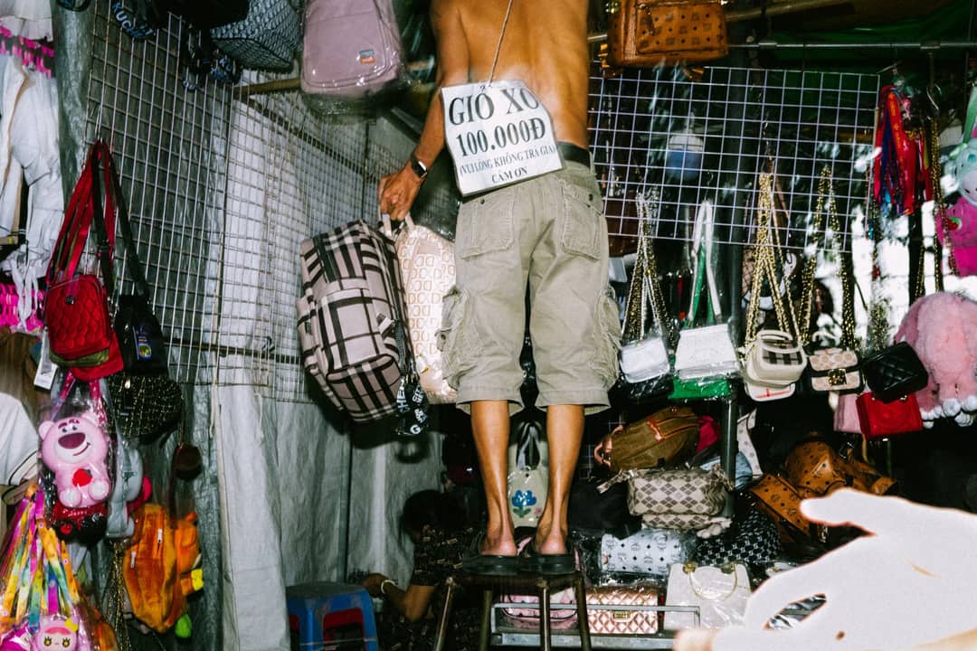 A street vendor selling assorted handbags at a bustling market stall, vibrant and busy.