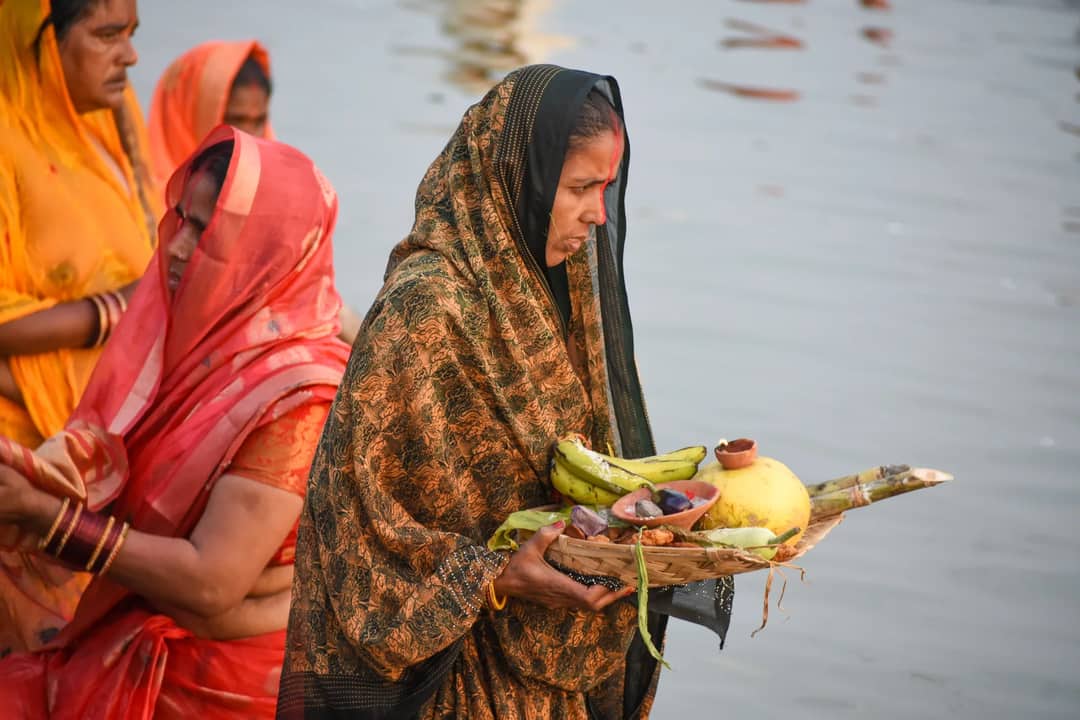 Women in traditional attire perform a religious ceremony at a river, holding offerings.