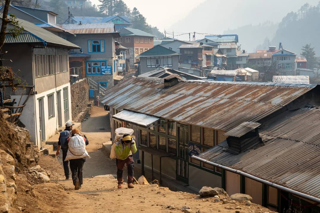 Villagers carrying loads on a rustic pathway in Himalayan village setting.