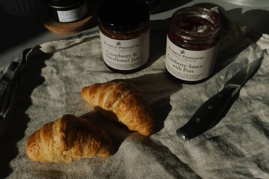 Still life of crispy tasty croissants with opened jam and sauce pots placed on gray fabric napkin near knife in rustic style