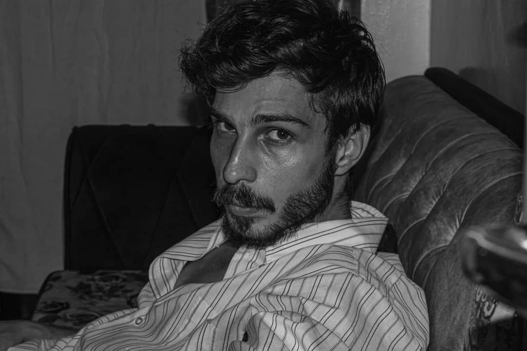 Moody black and white portrait of a bearded man in a striped shirt relaxing on a couch indoors.