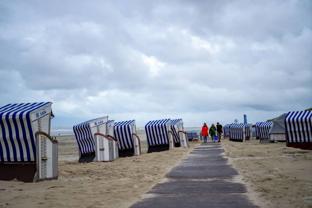 Four people stroll on a cloudy beach with striped huts, creating a calm seaside scene.