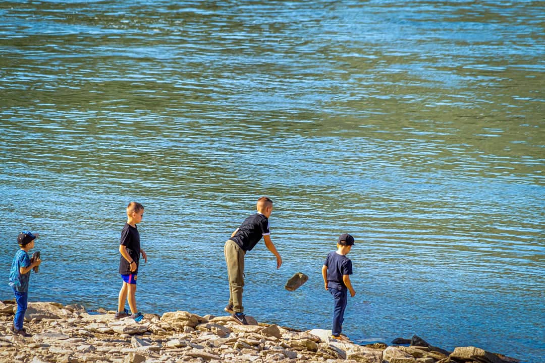 Four children enjoying skipping rocks by the riverbank on a sunny day. Perfect for depicting carefree childhood moments.