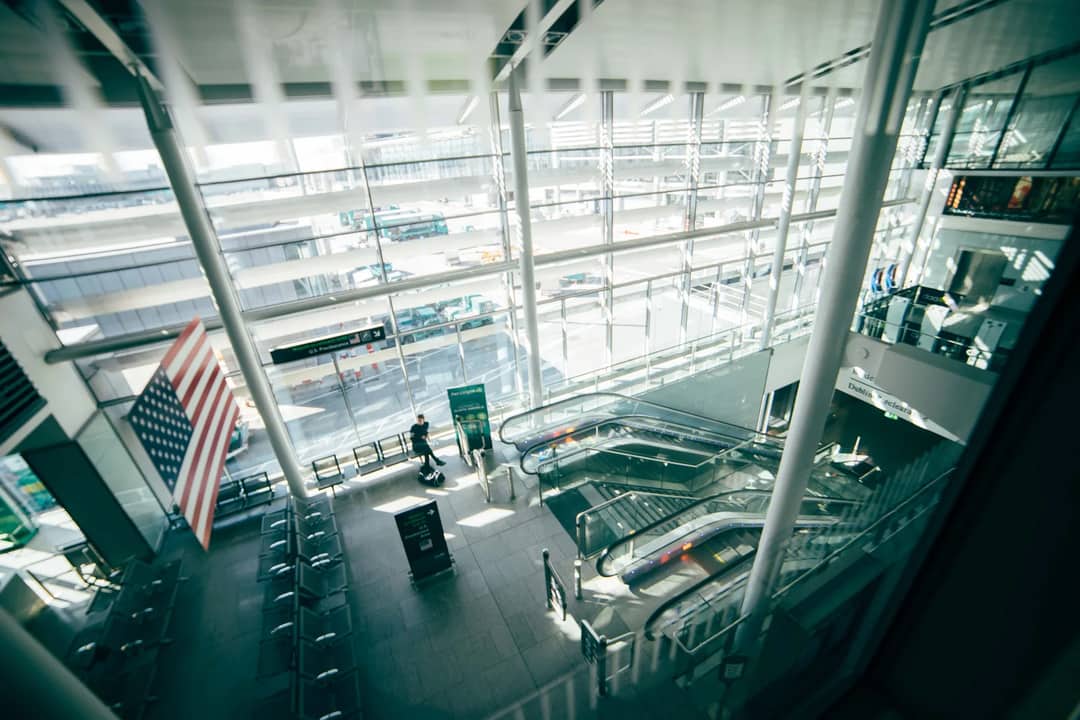 A bright, spacious airport terminal featuring expansive glass walls, escalators, and American flag.
