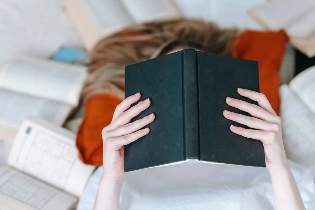 From above of unrecognizable female reading black book with hardcover while lying on floor near various literature in light room