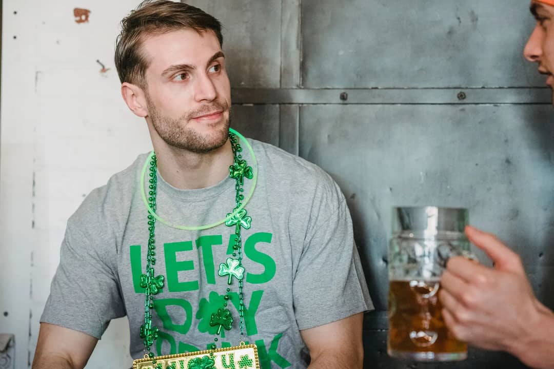 Positive bearded male wearing neon green glow stick necklace and necklace with shamrock and gray t shirt devoted to St Patrick Day communicating with friend in pub while celebrating feast