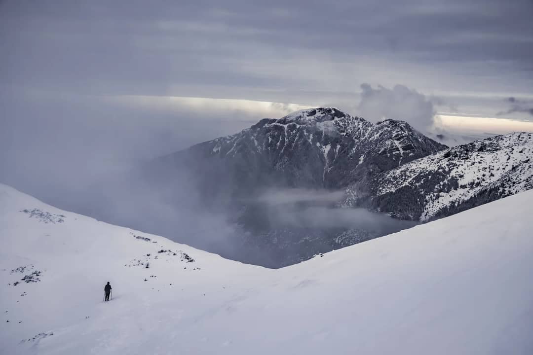 Lone hiker traversing a snow-covered mountain path with dramatic clouds.