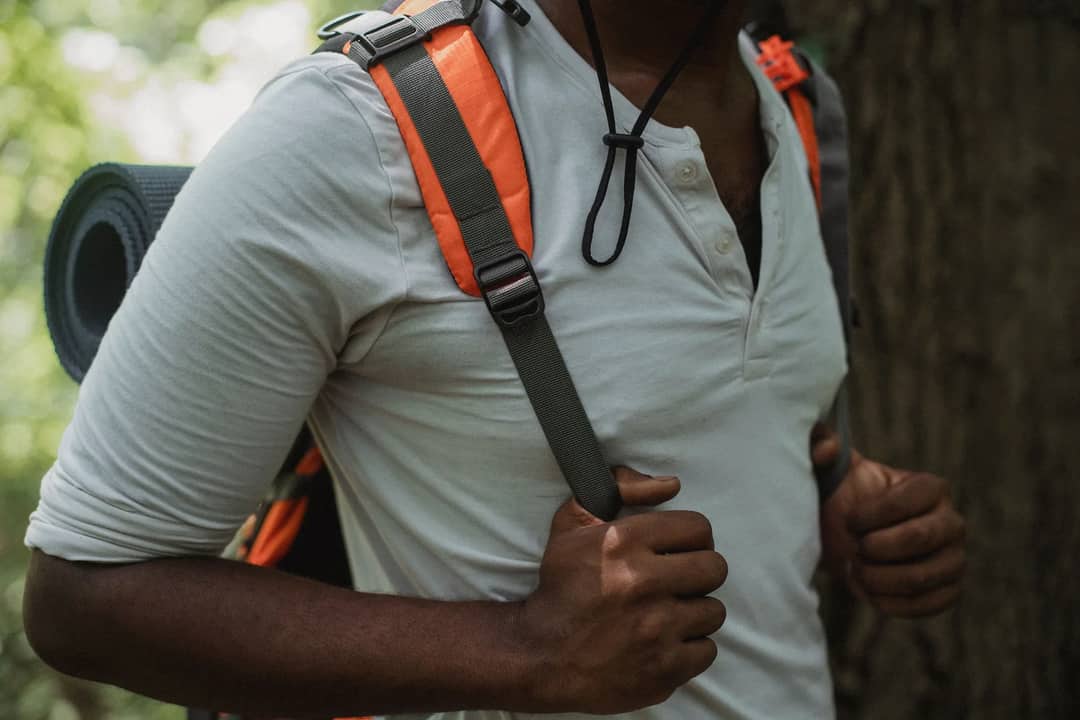 Crop anonymous ethnic male hiker with rucksack and rolled mat exploring summer forest during trip
