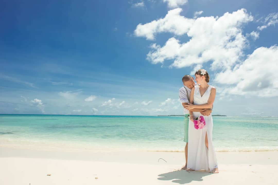A couple embraces romantically on a sunny beach by the ocean with clear skies.
