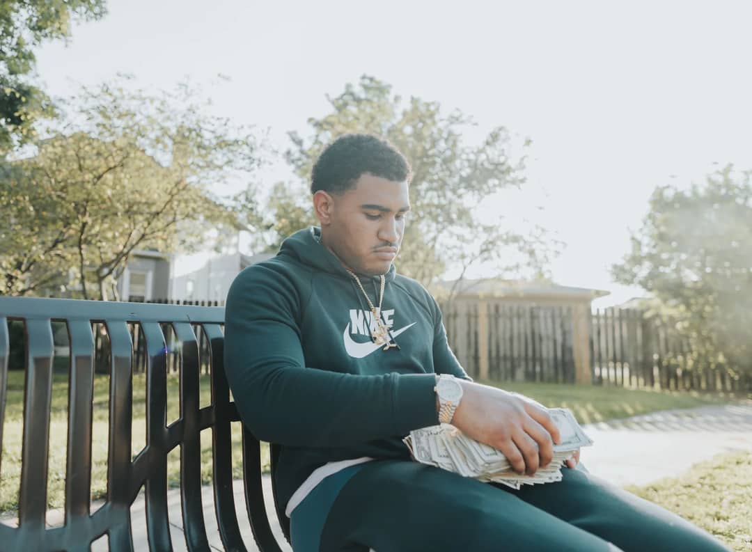 Young man in a hoodie counts stacks of cash on a park bench.