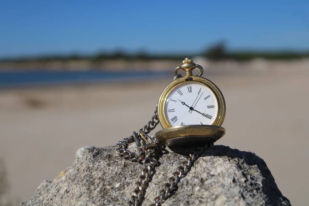 A classic pocket watch resting on a rock by the beach in El Puerto de Santa María, Spain, under clear skies.