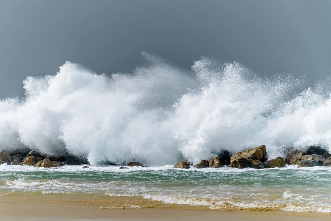 Big foamy waves splashing in stormy sea with stones near sandy wet shore under cloudless gray sky in gloomy day