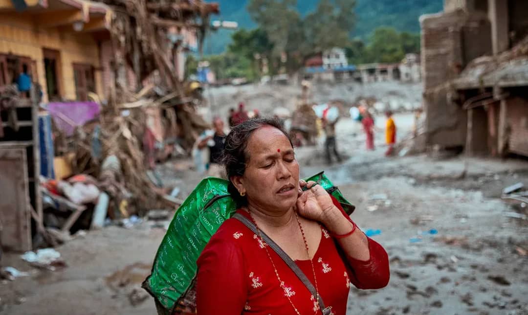 A woman carrying salvaged belongings in a green sack walks through a silt-covered street of damaged houses in the Bhote Koshi corridor after the 2026 flood, with other survivors picking through debris behind her