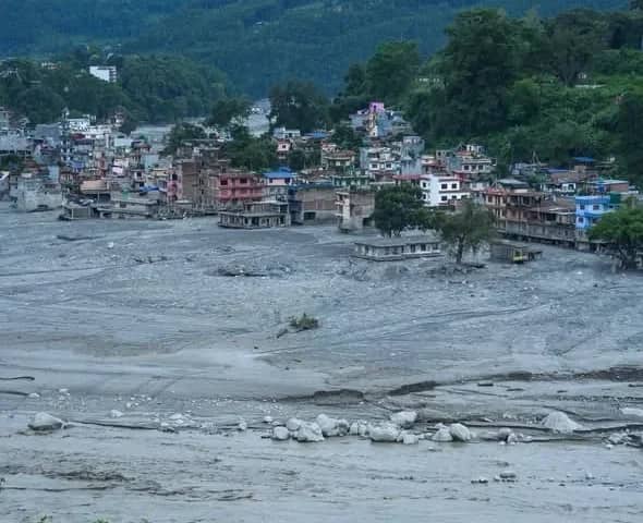 A hillside bazaar town with its lowest streets buried under a wide grey silt plain, the river still running brown below