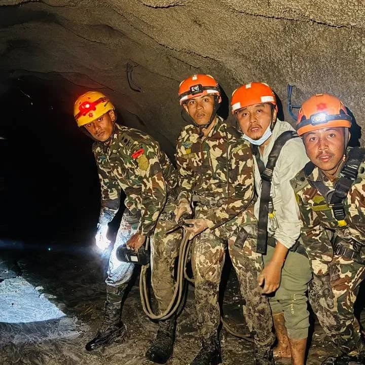 Four Nepal Army rescuers in camouflage and orange helmets with headlamps standing on the wet floor inside the dark Rasuwagadhi hydropower tunnel during the search for missing workers