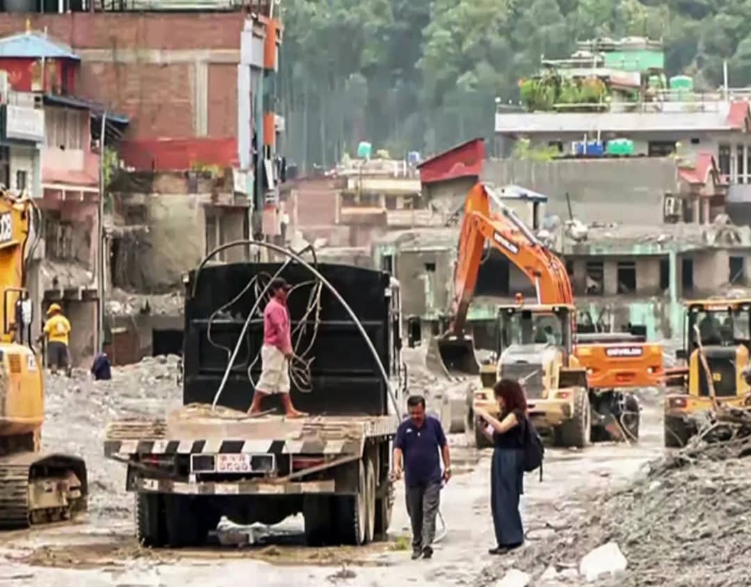 Excavators and a flatbed truck clearing grey flood debris from a buried street at Trishuli in Nuwakot, with gutted concrete buildings on one side and intact houses on the hillside above
