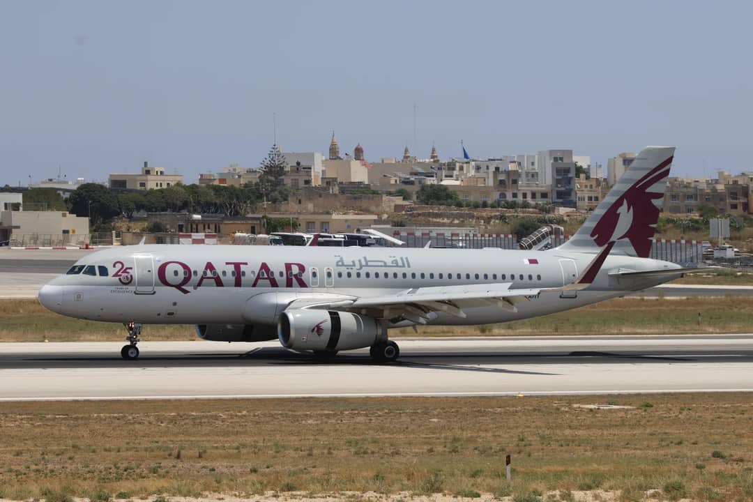 Qatar Airways Airbus A320 parked at the airport runway with urban background.