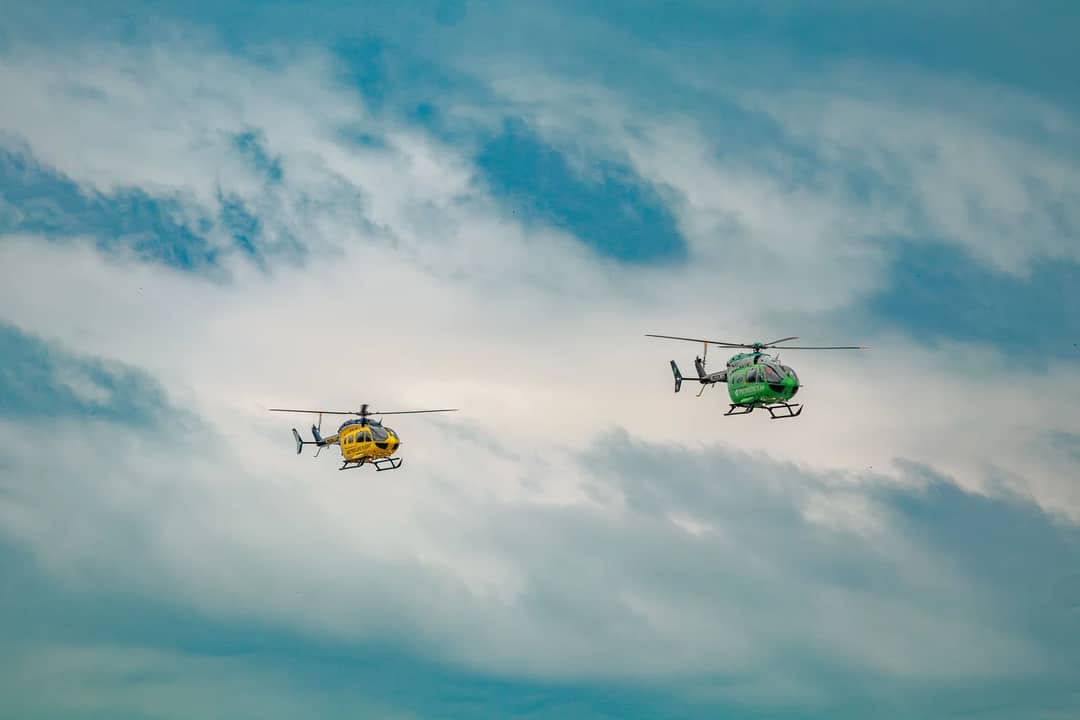 Two helicopters, one yellow and one green, soar through a bright cloudy sky.