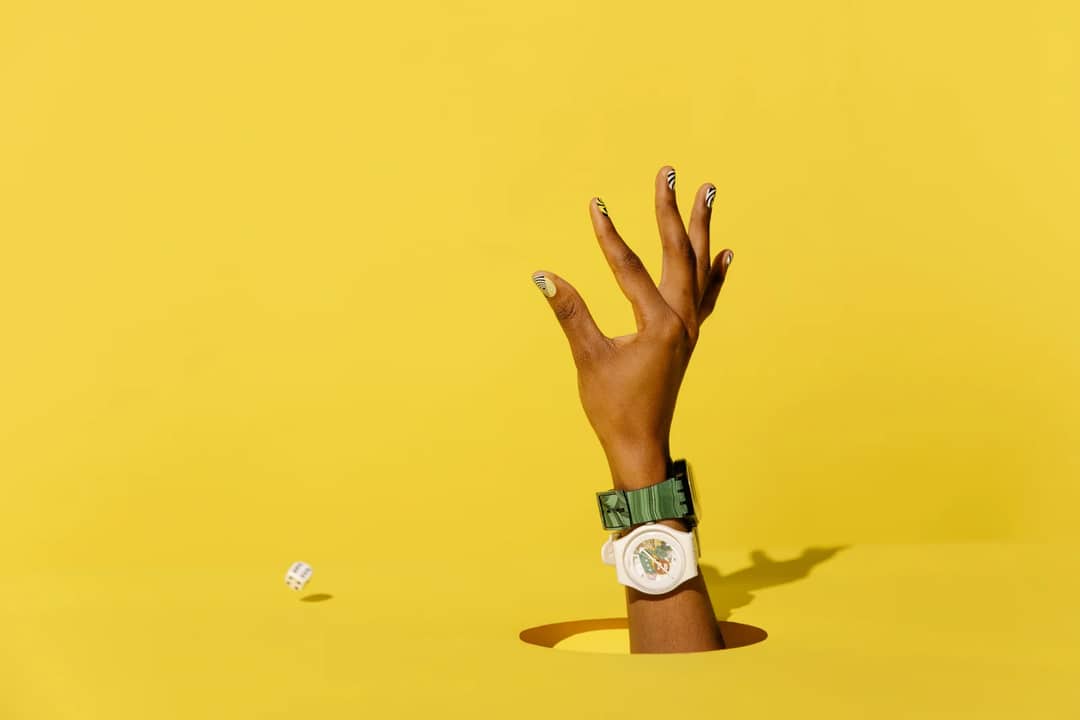 A hand wearing a fashionable watch extends through a yellow backdrop with a dice in motion.