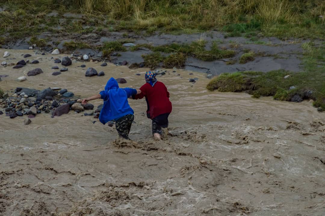 Two individuals navigating through a turbulent river crossing during a flood.
