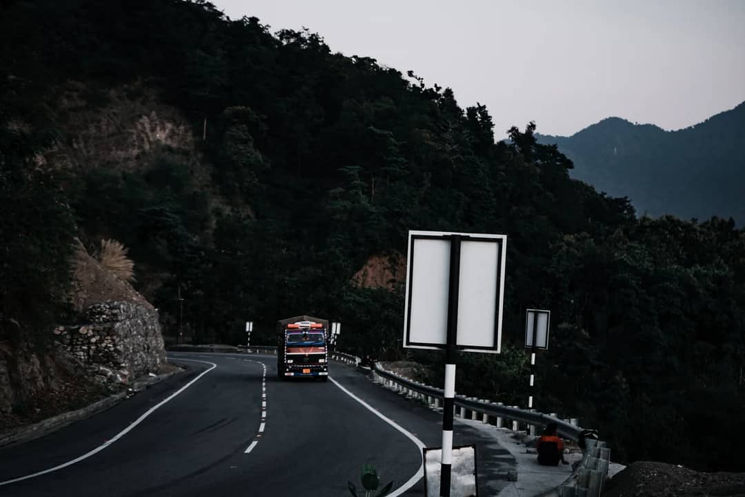 A truck navigates a winding mountain road surrounded by lush greenery and towering peaks.