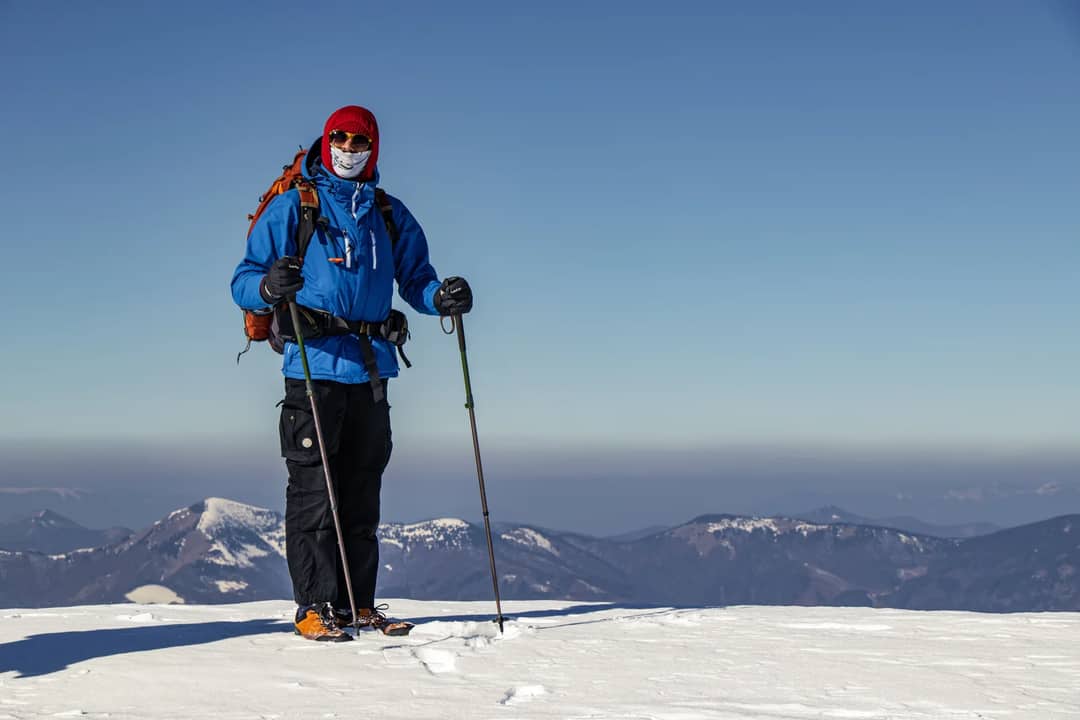 A lone hiker in winter gear stands on a snowy mountain summit with trekking poles.