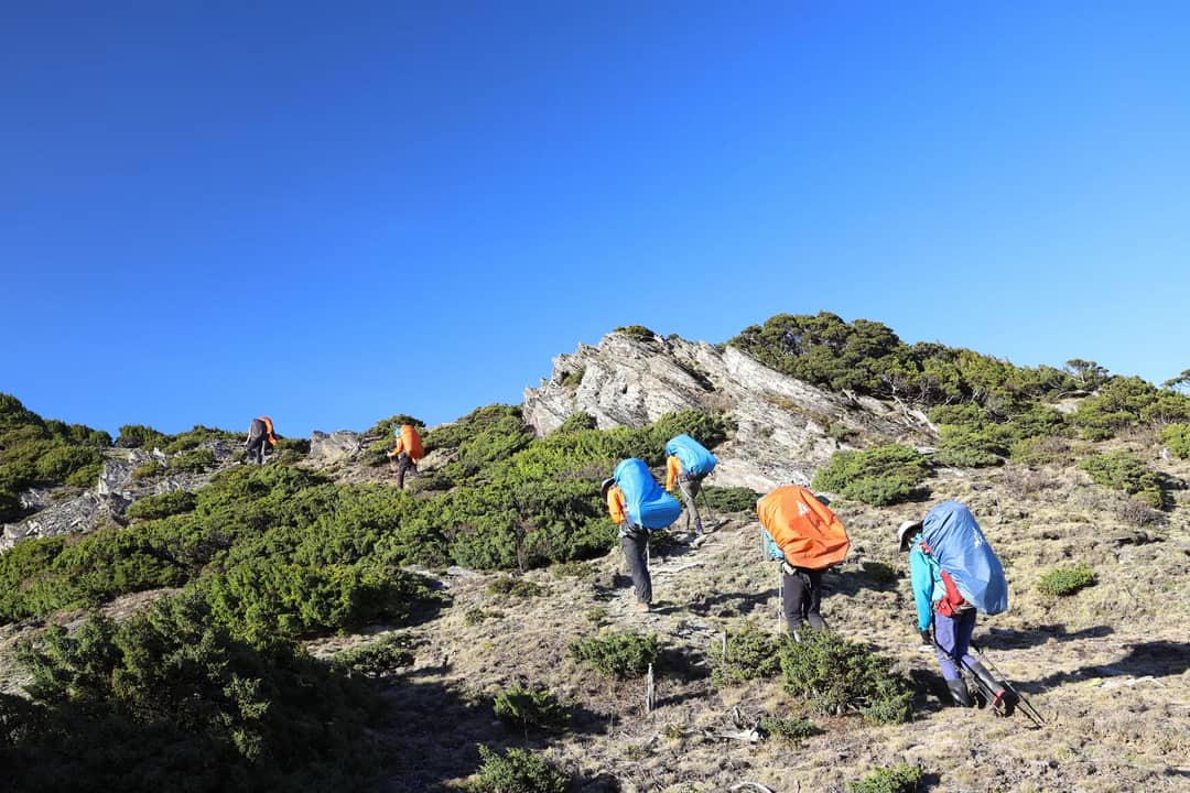 A group of hikers with backpacks trekking up a mountainous path in Taiwan under a clear blue sky.