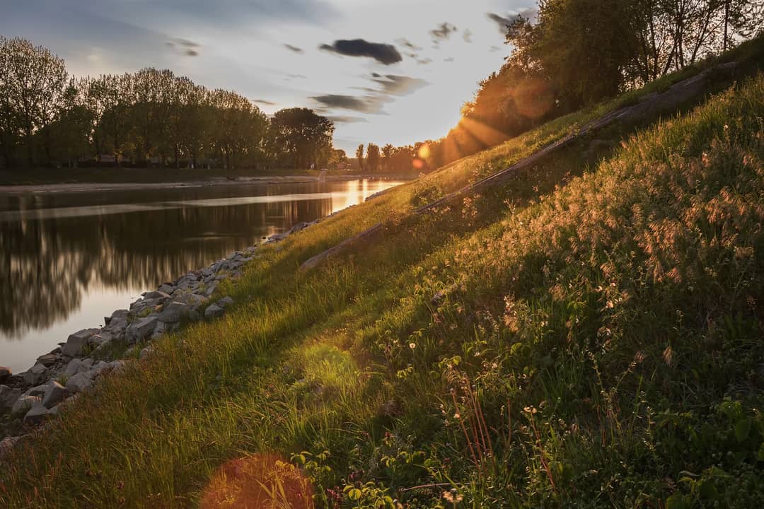 A peaceful sunset scene by a river, with sunlight illuminating the grassy slope and trees in the distance.