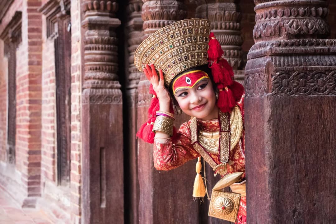 A young girl in traditional Nepalese dress peeks through ornate wooden architecture.