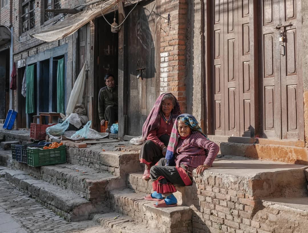 Two elderly women sit outside their home in Bhaktapur, Nepal, enjoying sunlight.
