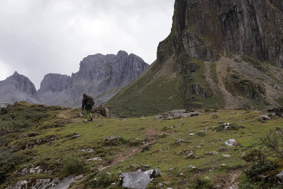 A lone hiker walks up a rocky trail towards towering mountains under a cloudy sky.