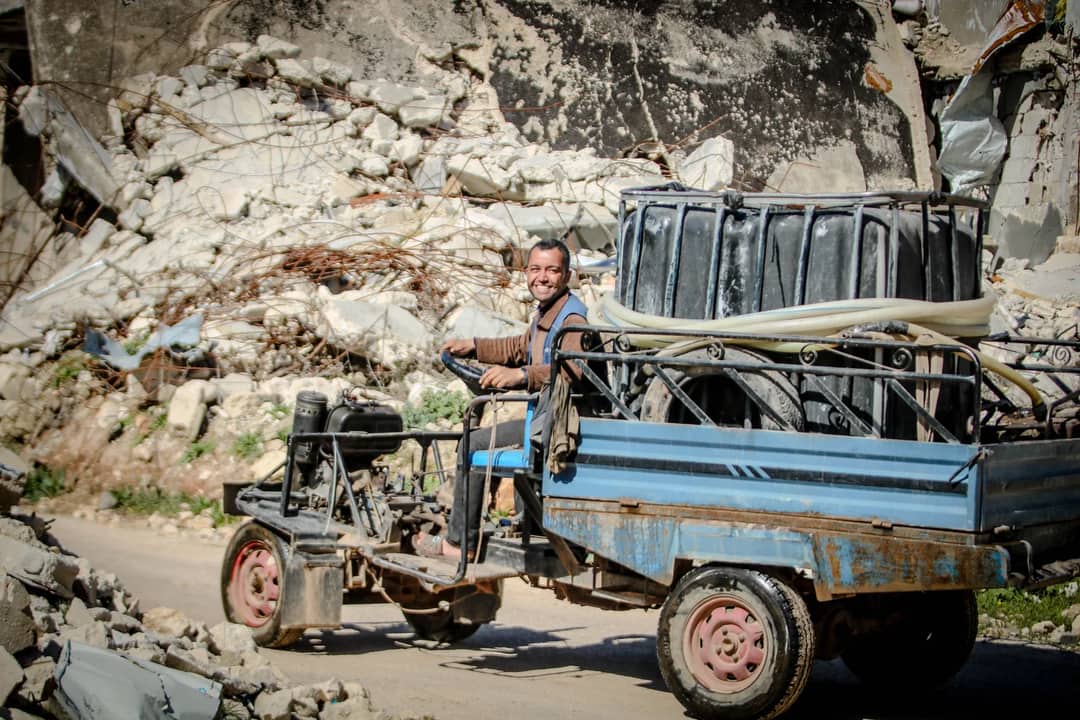 Smiling man driving a makeshift vehicle through rubble-strewn street in Idlib, Syria under bright sunlight.