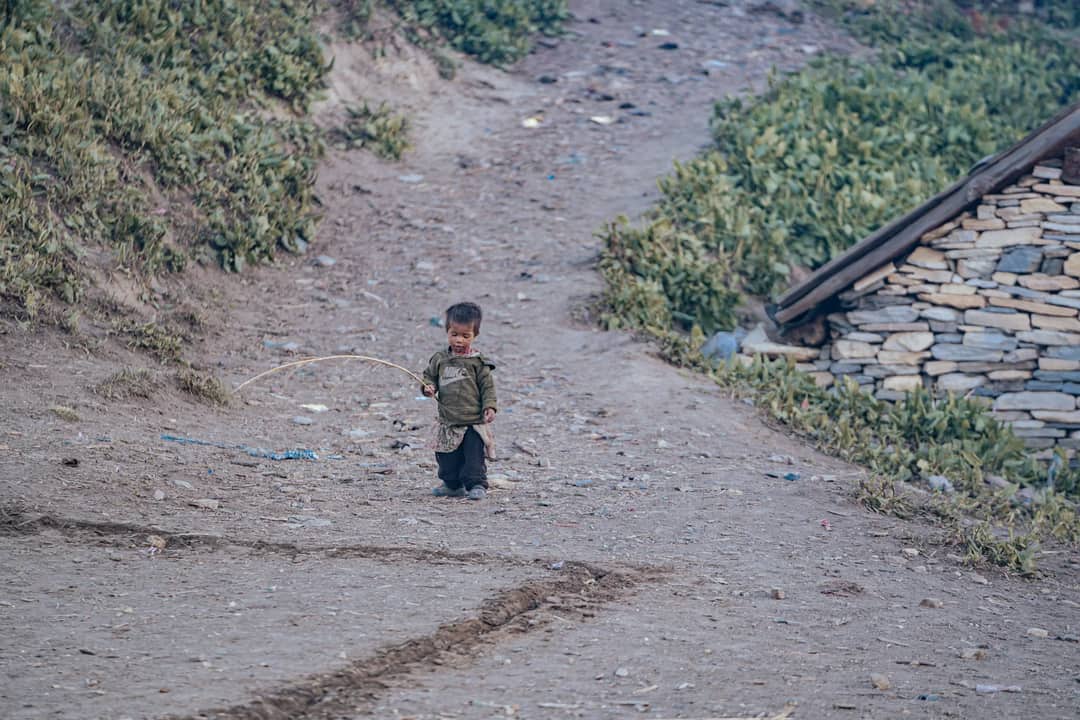Young child playing with a stick on a dirt path in a rural setting. Captured in daylight.