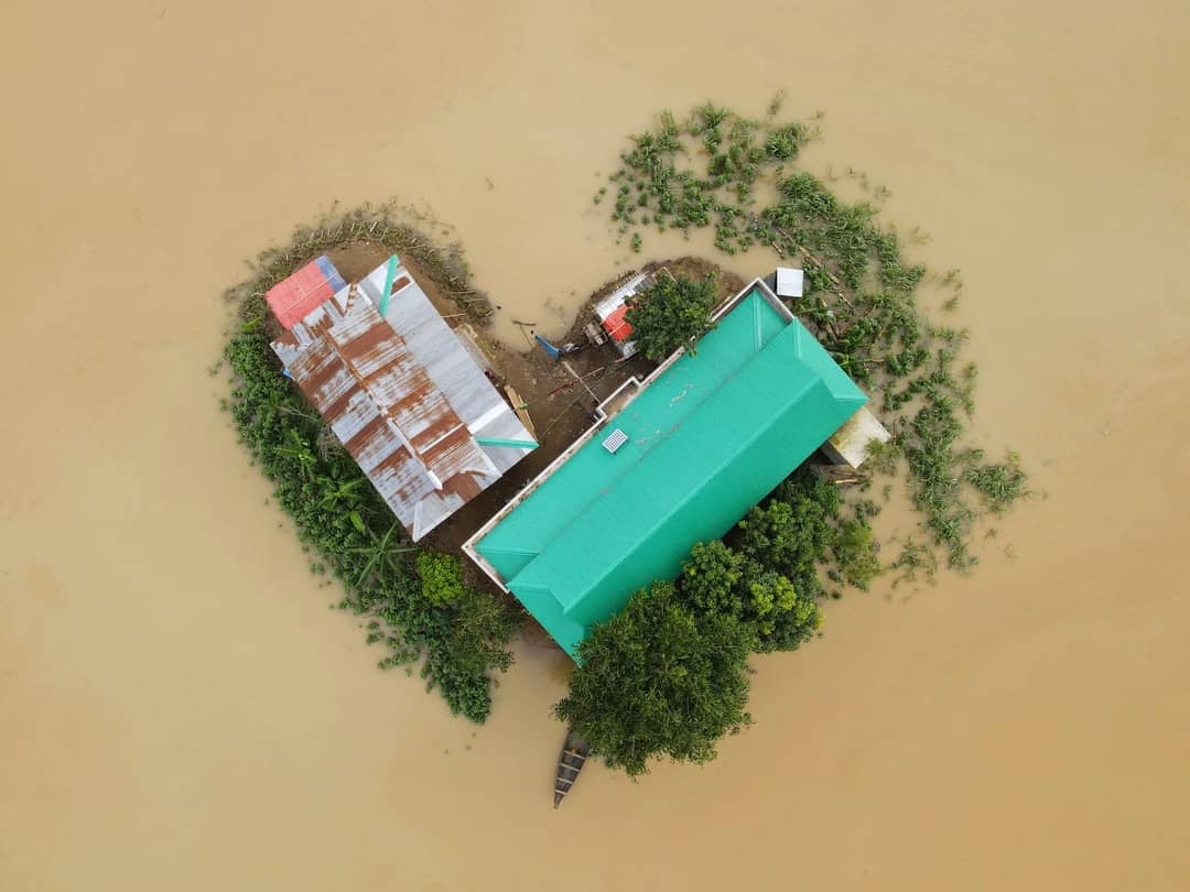 Top-down view of a flood-affected area in Bangladesh showing buildings and surrounding submerged land.